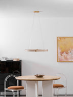 A slim travertine ring pendant from Clérachic hangs in front of a white wall above a round table with a dark bowl and clear chairs.
