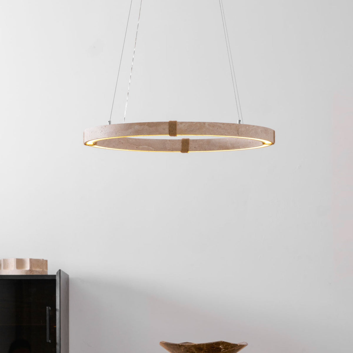 Lit travertine ring pendant from Clérachic hung low in a white room, with a black cabinet and a dark bowl on a table below.