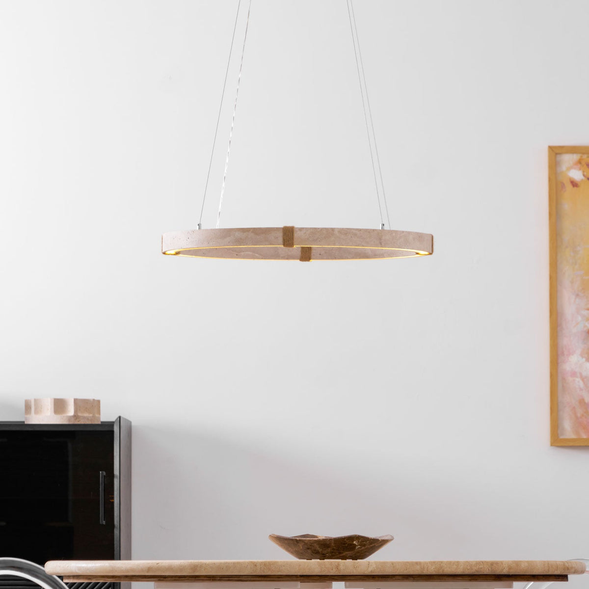 A Clérachic travertine ring pendant glows warm under its stone ring, hung in front of a painting with a black cabinet below.