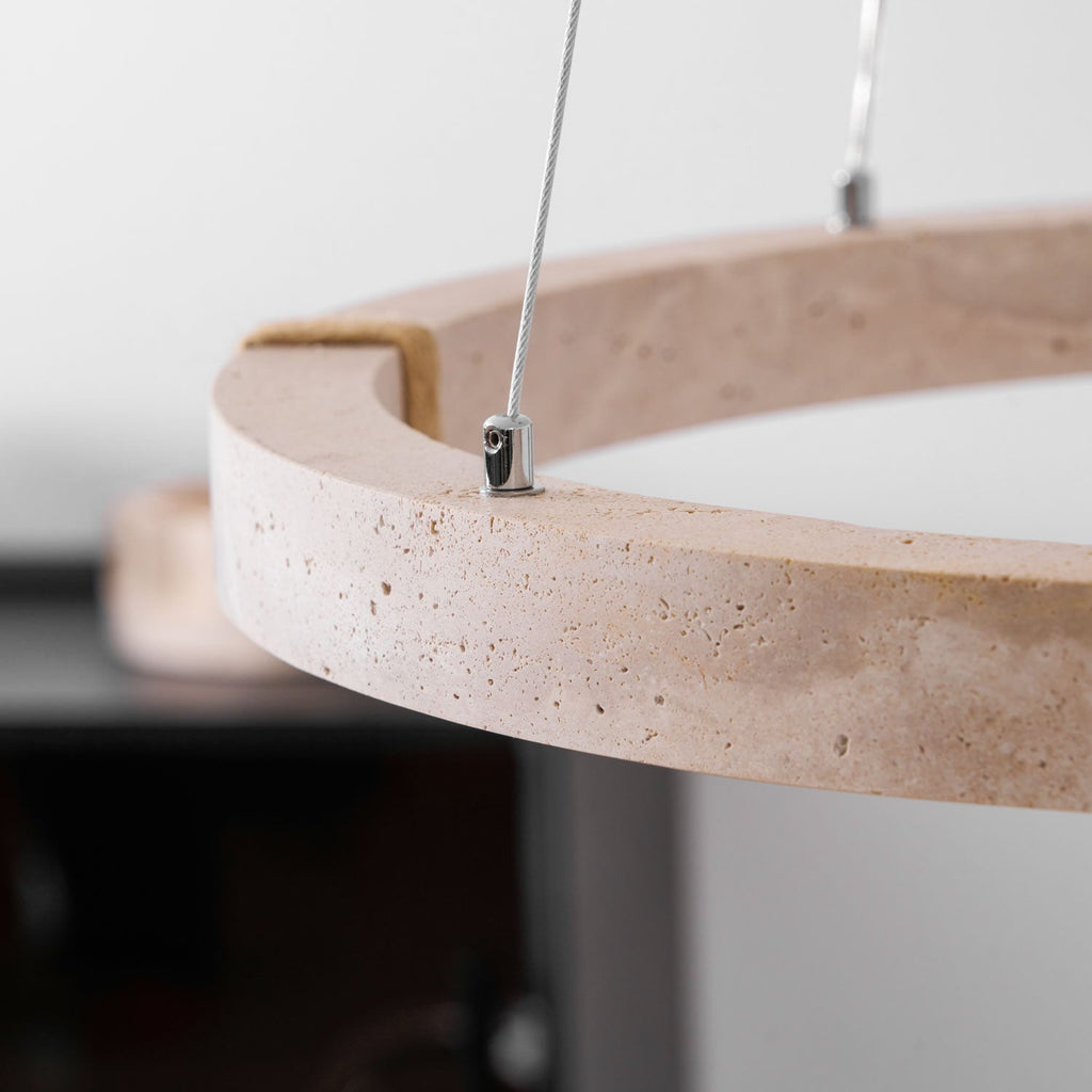 Close view of the pitted travertine edge and the steel cable fixing on this Clérachic ring pendant, with a dark cabinet blurred behind.