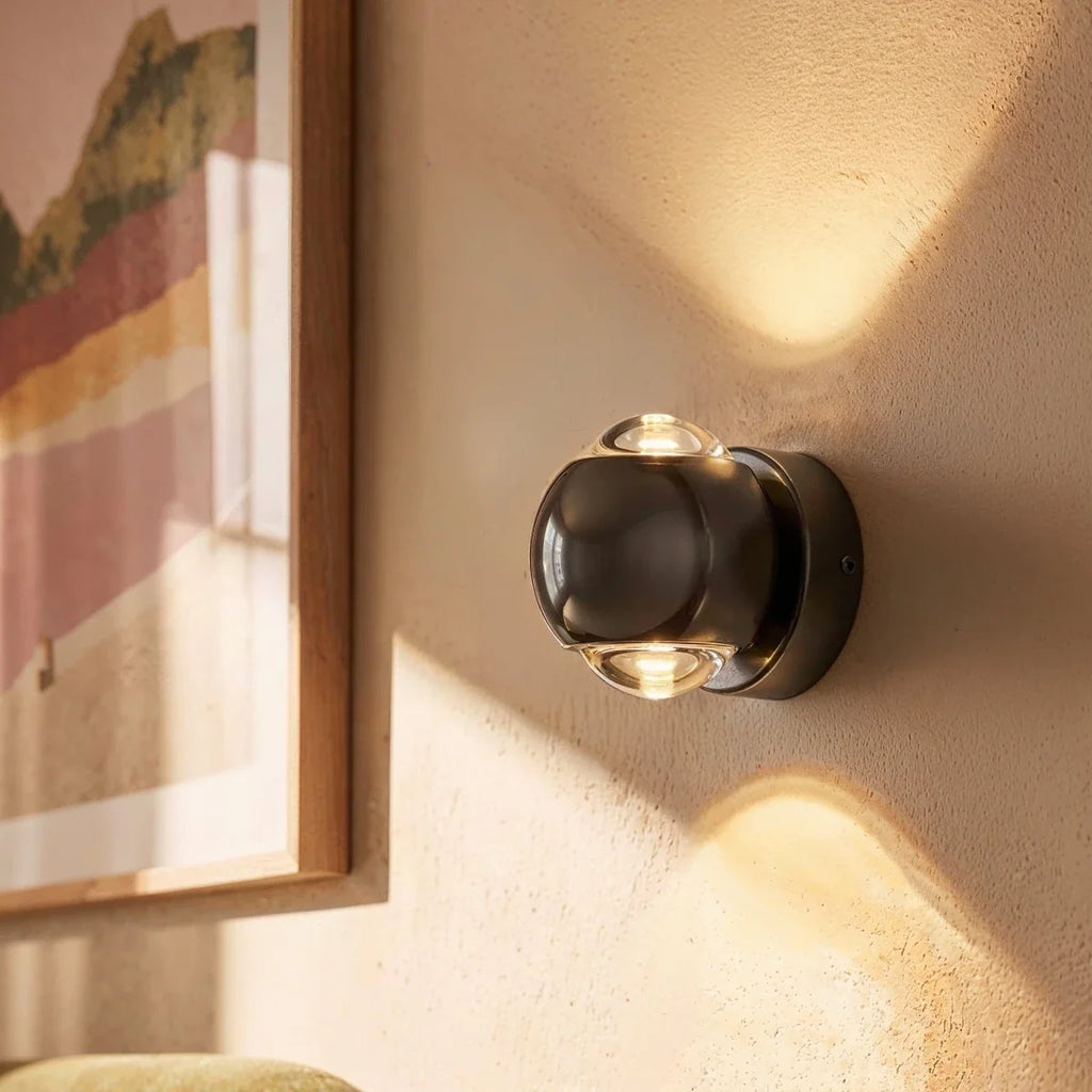 Close view of a Clérachic dark round wall sconce with clear lenses, beside a framed print, lighting the wall up and down