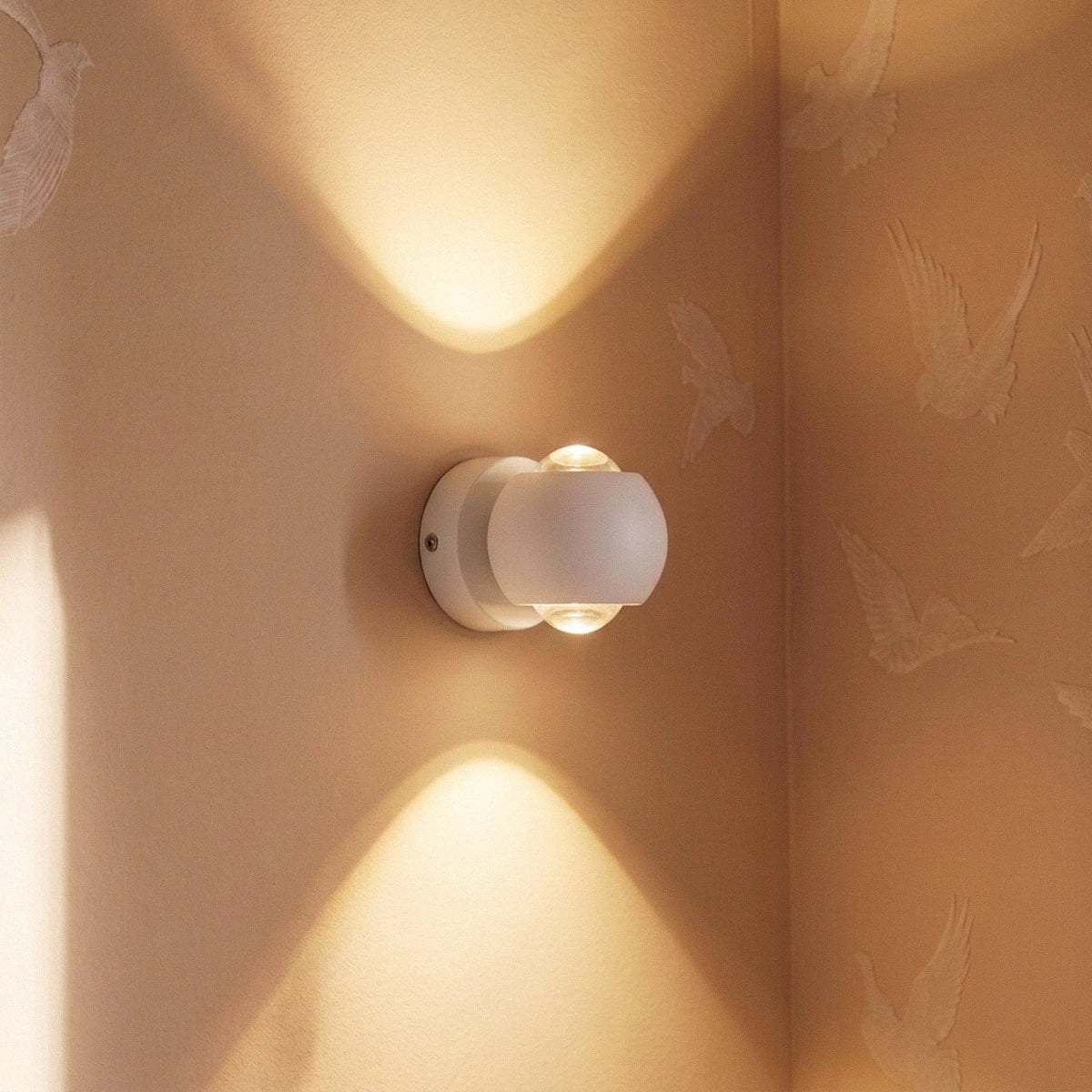 Clérachic white round wall sconce glowing up and down a wall corner with a soft bird-motif wallpaper