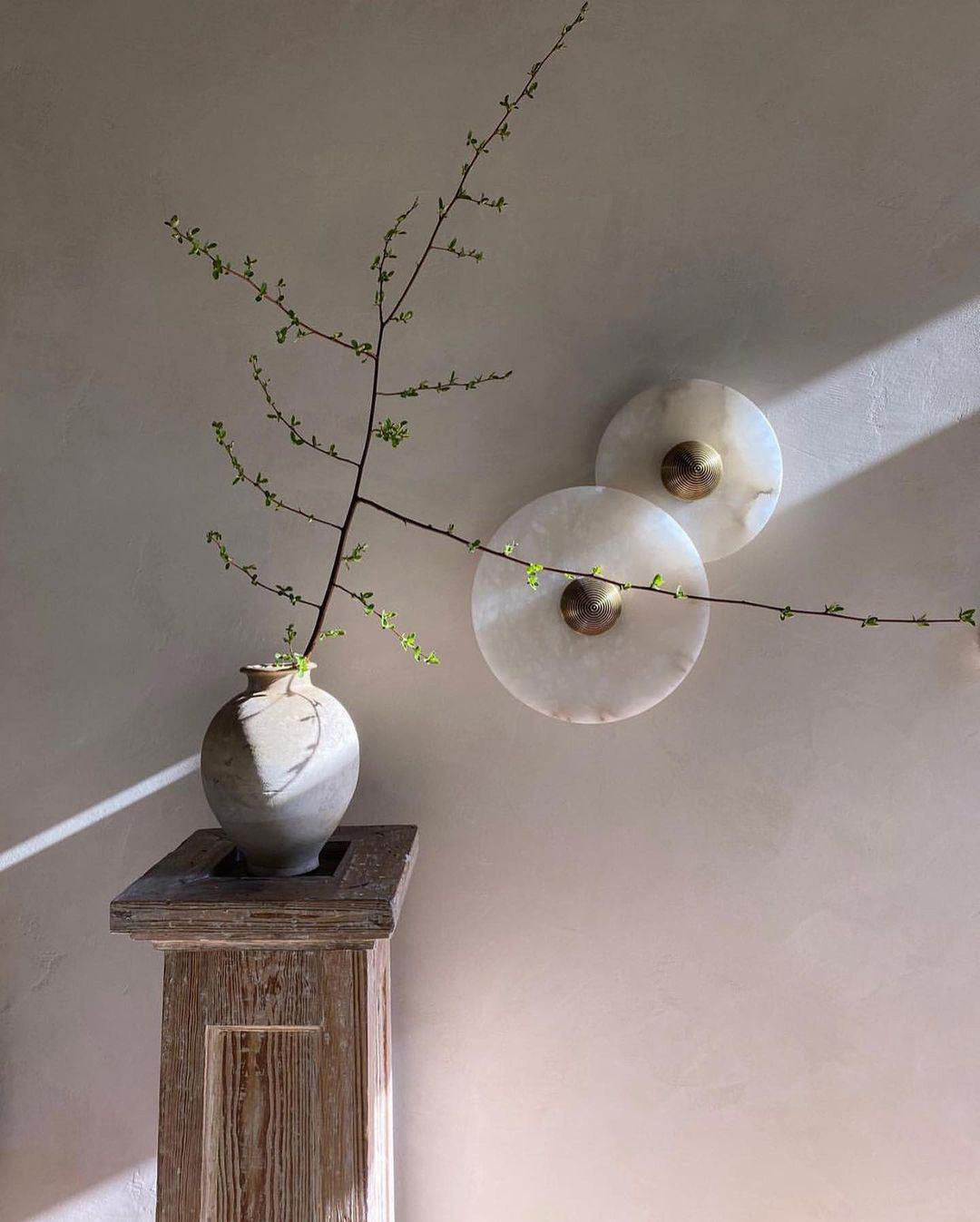 Two Clérachic round alabaster sconces overlapping on a pale wall behind a stoneware vase.