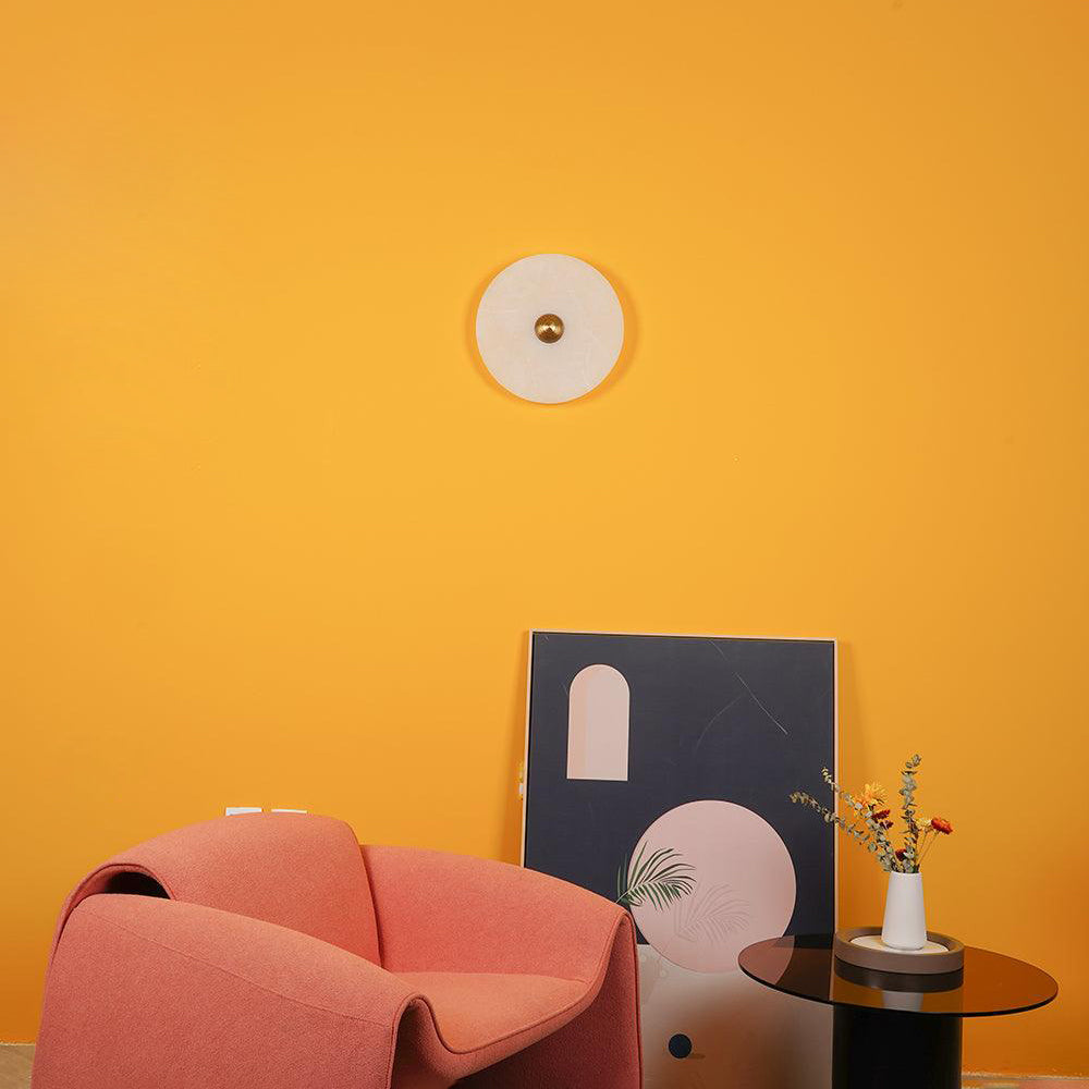A single Clérachic round alabaster sconce on a bright yellow wall above a coral armchair.