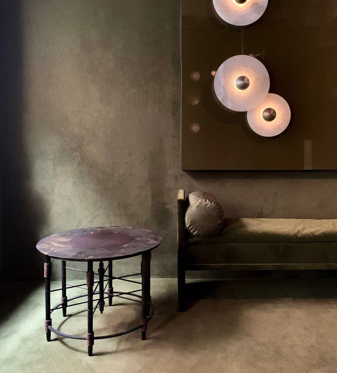 Clérachic round alabaster sconces lit in a dim olive room above a low bench and table.