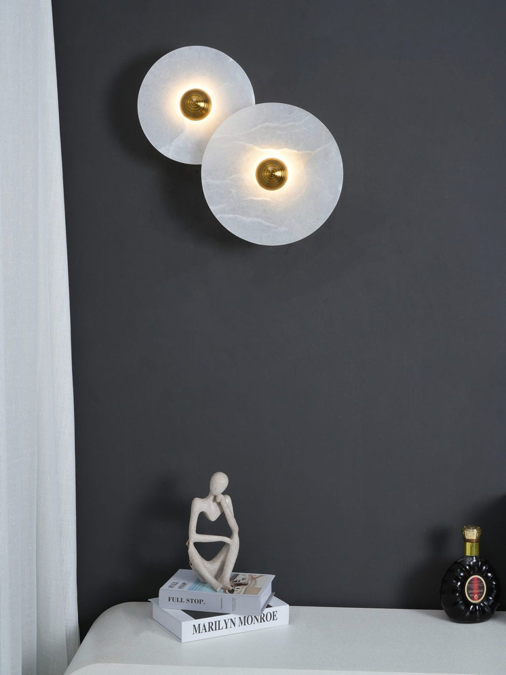 Clérachic round alabaster sconces mounted on a charcoal wall above books and a figurine.