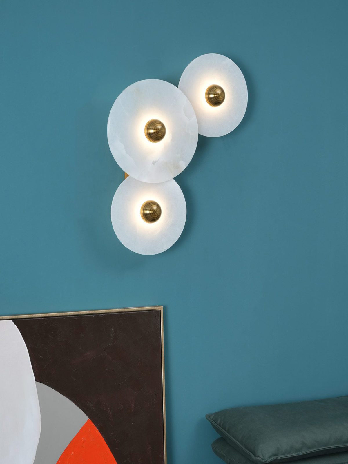 Three Clérachic round alabaster sconces lit on a deep teal wall above stacked artwork.