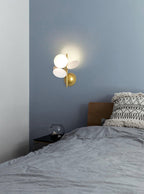 Small lit Clérachic wall light on a blue-grey bedroom wall above a bed with grey linen and a patterned cushion.