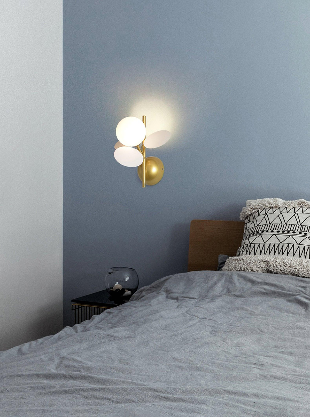 Small lit Clérachic wall light on a blue-grey bedroom wall above a bed with grey linen and a patterned cushion.