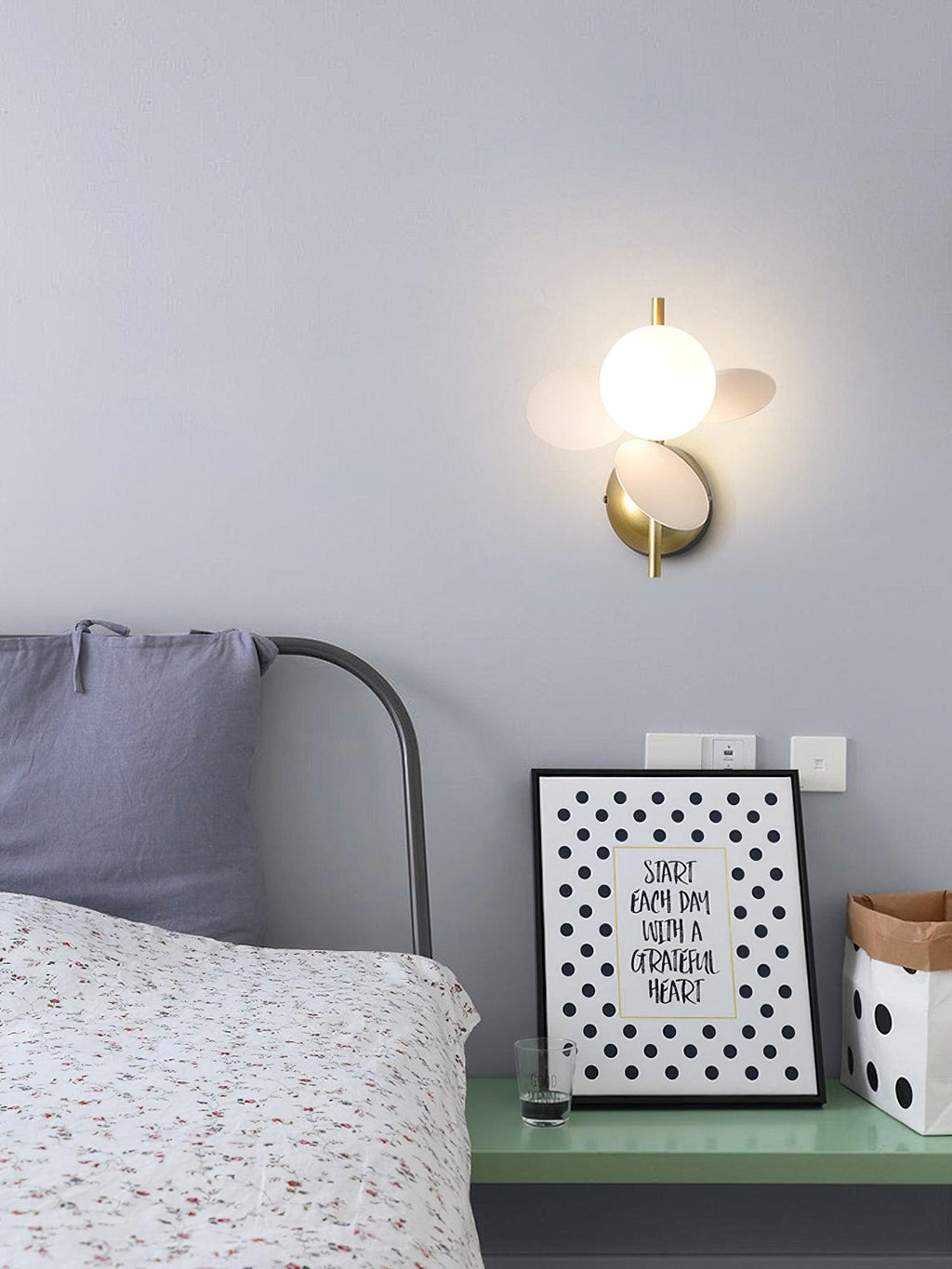 White Clérachic wall light glowing above a bedside shelf with a framed print, in a bedroom with a pale lilac-grey wall.