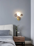 Grey-disc Clérachic wall light lit on a blue-grey wall above a wooden bedside table with a small vase of stems.