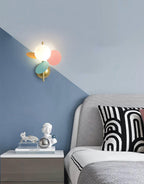 Multicolour Clérachic wall light on a two-tone blue wall above a bedside table with small ornaments and a patterned cushion.