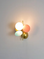 Close-framed Clérachic wall light on a soft grey wall, its lit globe casting a warm glow past the pink and mint discs.