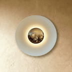Clérachic round wall lamp in studio light: a pale white disc with a mirrored brass dome and a soft glow around it.