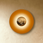 Clérachic round wall lamp in studio light with an amber orange disc and a brass mirror dome surrounded by a warm glow.