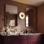 Clérachic round wall lamp with a mirrored brass dome, hung between two gold-framed mirrors above a marble-topped vanity unit.
