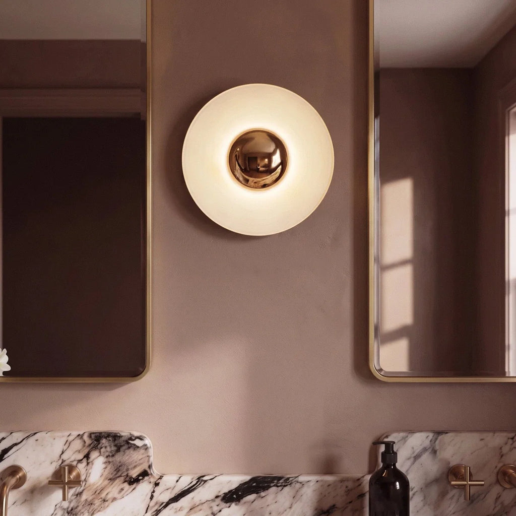 Close up of the Clérachic round wall lamp glowing on a dusty rose wall, flanked by gold-framed mirrors and brass taps.
