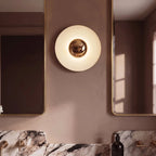 Close up of the Clérachic round wall lamp glowing on a dusty rose wall, flanked by gold-framed mirrors and brass taps.