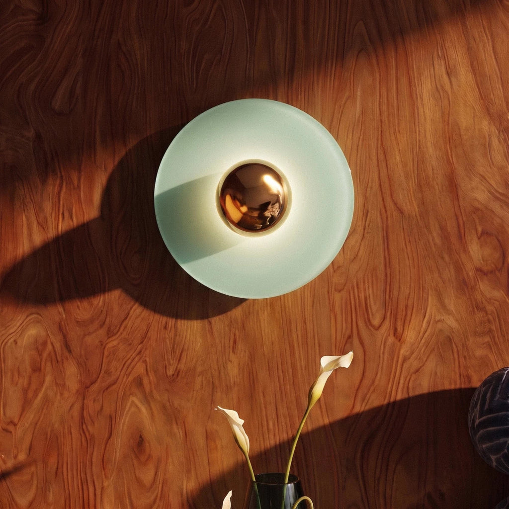 Clérachic round wall lamp with a pale green disc and brass mirror dome, casting a long shadow on walnut wood panelling in a room.