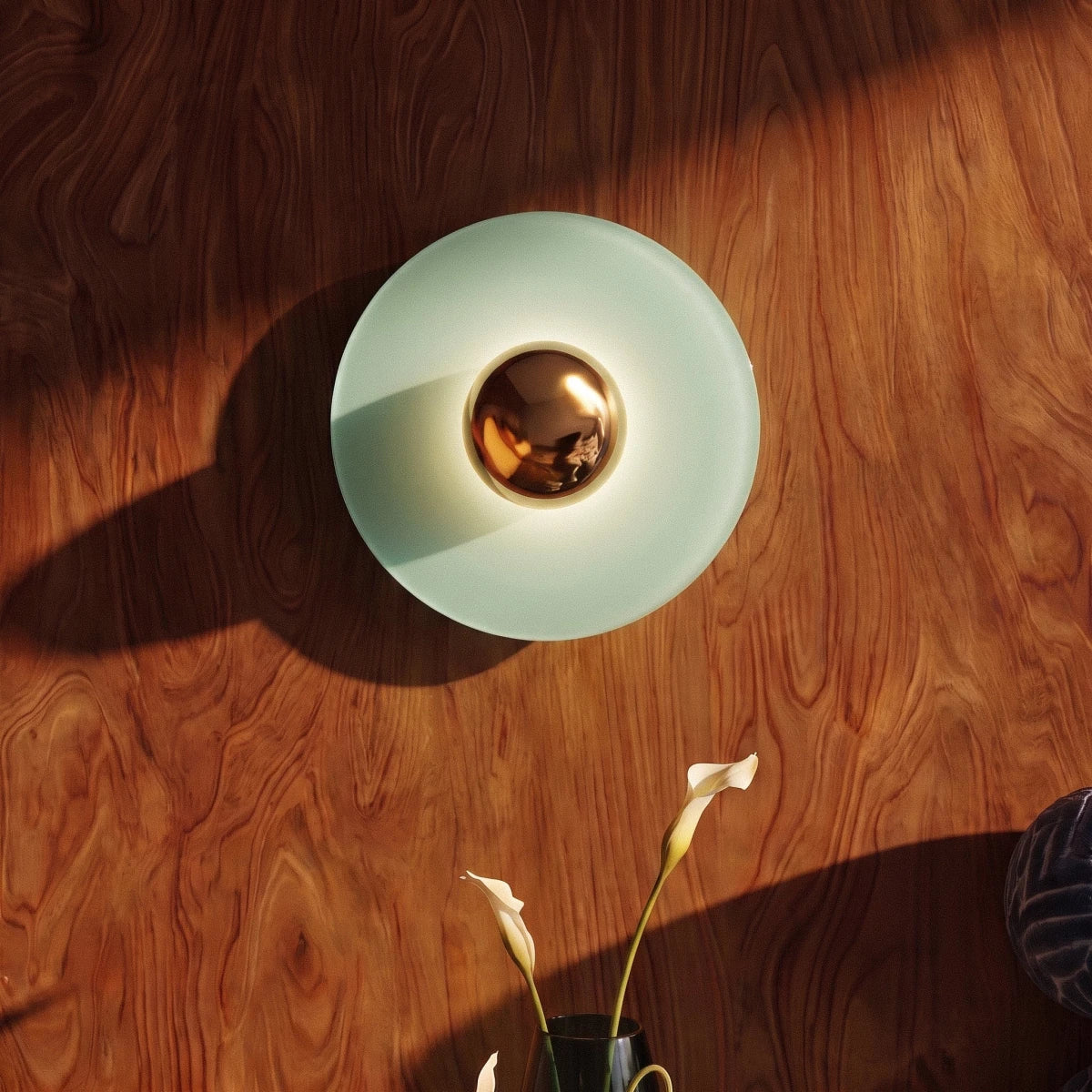 Clérachic round wall lamp with a pale green disc and brass mirror dome, casting a long shadow on walnut wood panelling in a room.