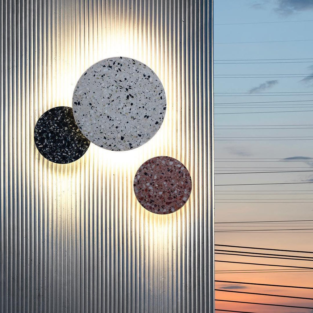 Three Clérachic round wall lights in terrazzo, black, white and pink, glow against a ribbed metal wall at sunset.
