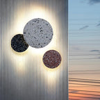 Three Clérachic round wall lights in terrazzo, black, white and pink, glow against a ribbed metal wall at sunset.