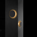 This Clérachic round wall light in plain black glows with a warm halo on a dark wall, with a second light visible beside a door.