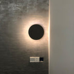 A plain black disc Clérachic wall light throws a warm halo of light across a rough dark wall, above a wall socket.