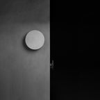 A Clérachic round wall light with a pale cement-grey face on a dark grey wall, switched off beside a black doorway.