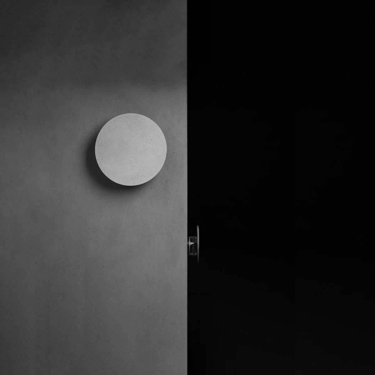 A Clérachic round wall light with a pale cement-grey face on a dark grey wall, switched off beside a black doorway.