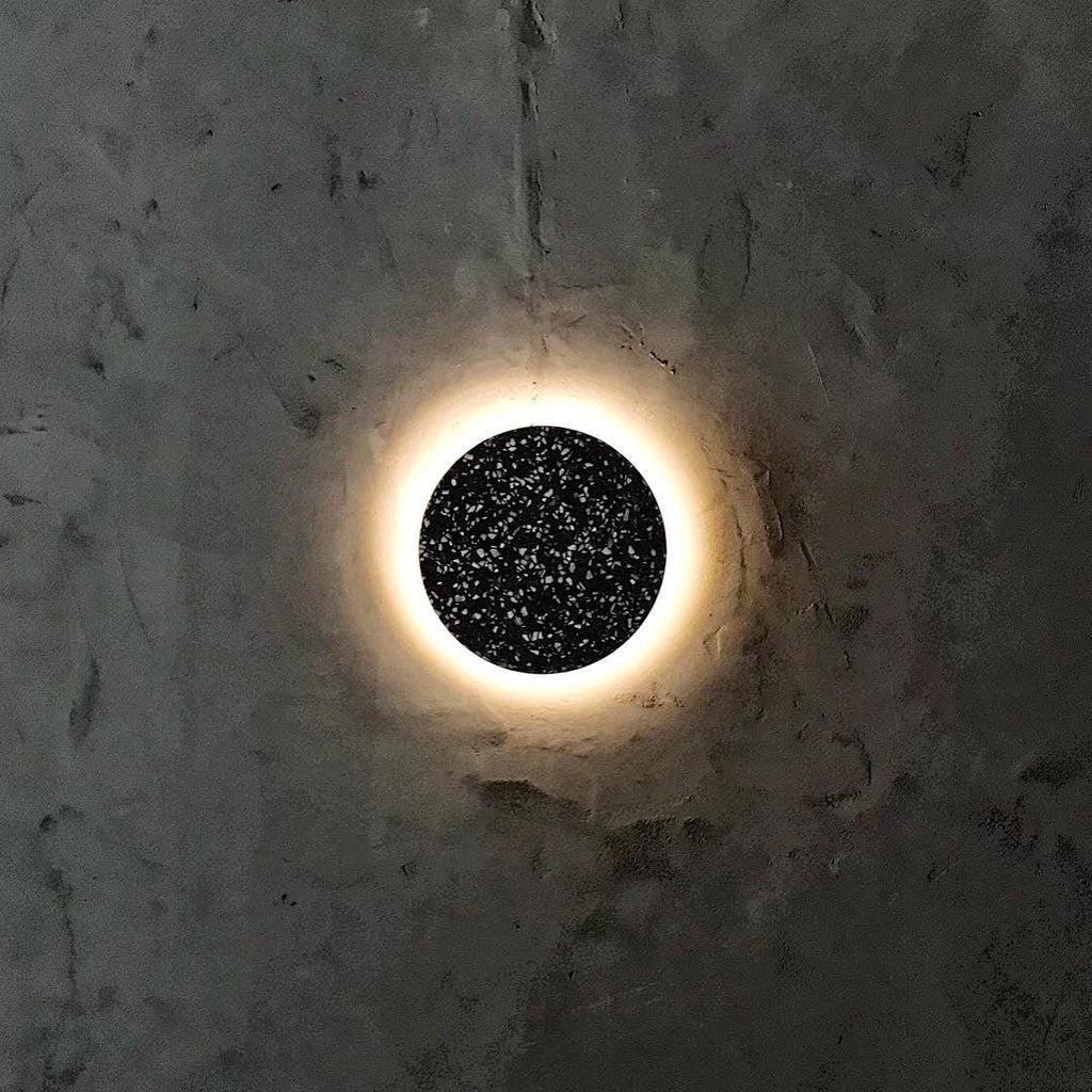 This Clérachic round wall light has a black terrazzo face and a warm ring of light on a rough dark plaster wall.