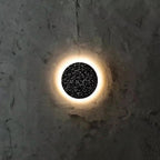 This Clérachic round wall light has a black terrazzo face and a warm ring of light on a rough dark plaster wall.