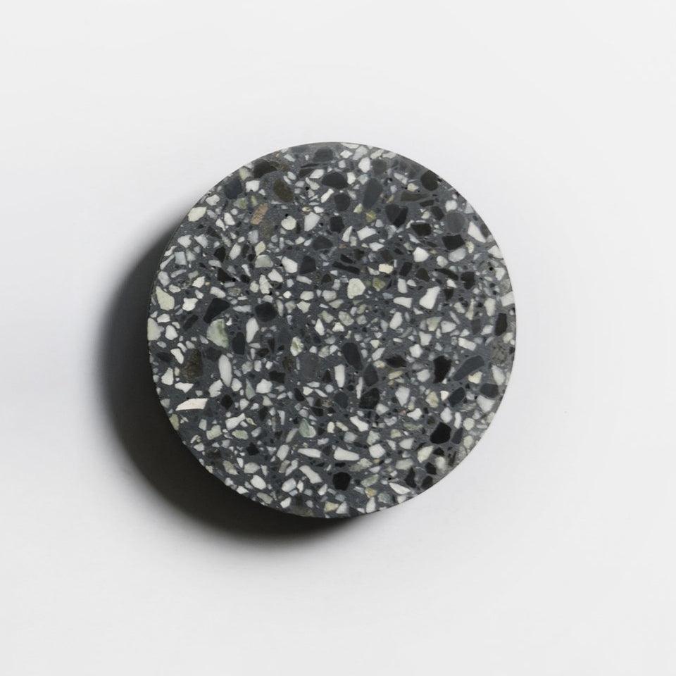 A Clérachic round wall light with a black terrazzo face and white and grey chips, casting a soft shadow on a pale wall.