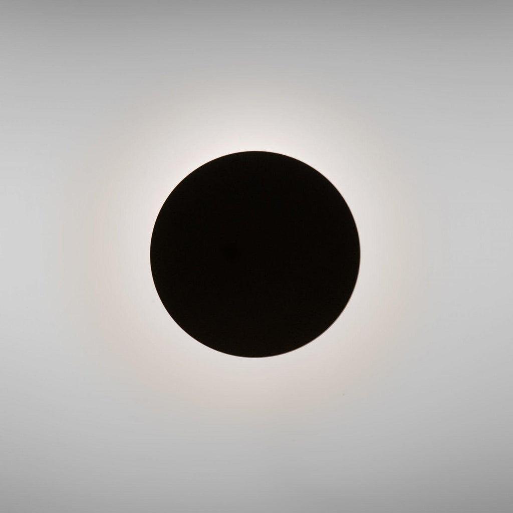 A plain black Clérachic disc wall light seen head-on, with a soft halo of warm light around its edge on a grey wall.