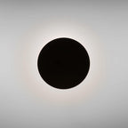 A plain black Clérachic disc wall light seen head-on, with a soft halo of warm light around its edge on a grey wall.