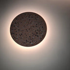 This Clérachic round wall light has a dark terrazzo face and a soft glow behind it on a plain grey wall.