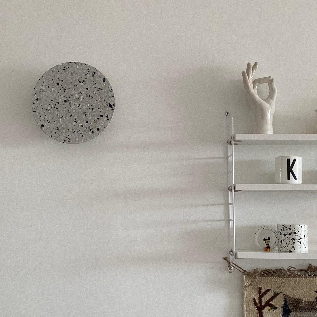 A grey terrazzo Clérachic round wall light hangs on a white wall beside a slim white shelf with a cup and a hand sculpture.