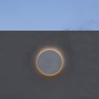 A pale Clérachic round wall light with a glowing halo, on a grey wall under a blue sky.