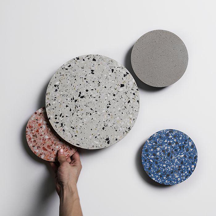 A hand holds a white terrazzo Clérachic wall light, with pink, blue and cement-grey discs on the wall beside it.