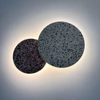Two overlapping Clérachic round wall lights, one dark red and one grey terrazzo, glow with a halo on a pale wall.