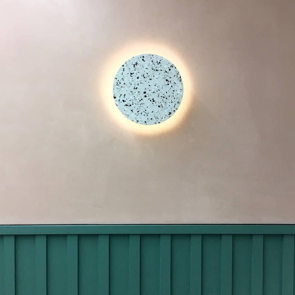 A pale blue-grey terrazzo Clérachic round wall light glows on a cream wall, above a green wooden panel.