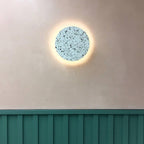 A pale blue-grey terrazzo Clérachic round wall light glows on a cream wall, above a green wooden panel.