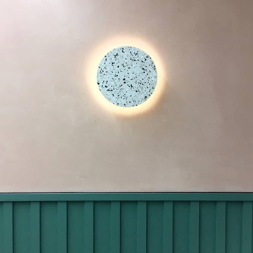 A pale blue-grey terrazzo Clérachic round wall light glows on a cream wall, above a green wooden panel.