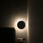 A plain black Clérachic round wall light glows beside a door in a dark bedroom, with a bed headboard at the left.