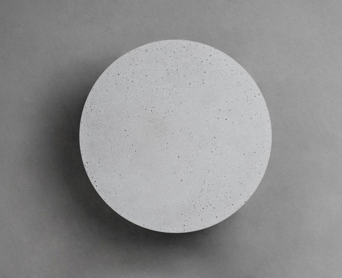 A Clérachic round wall light with a pale cement-grey face on a mid-grey wall, with a soft shadow at its edge.