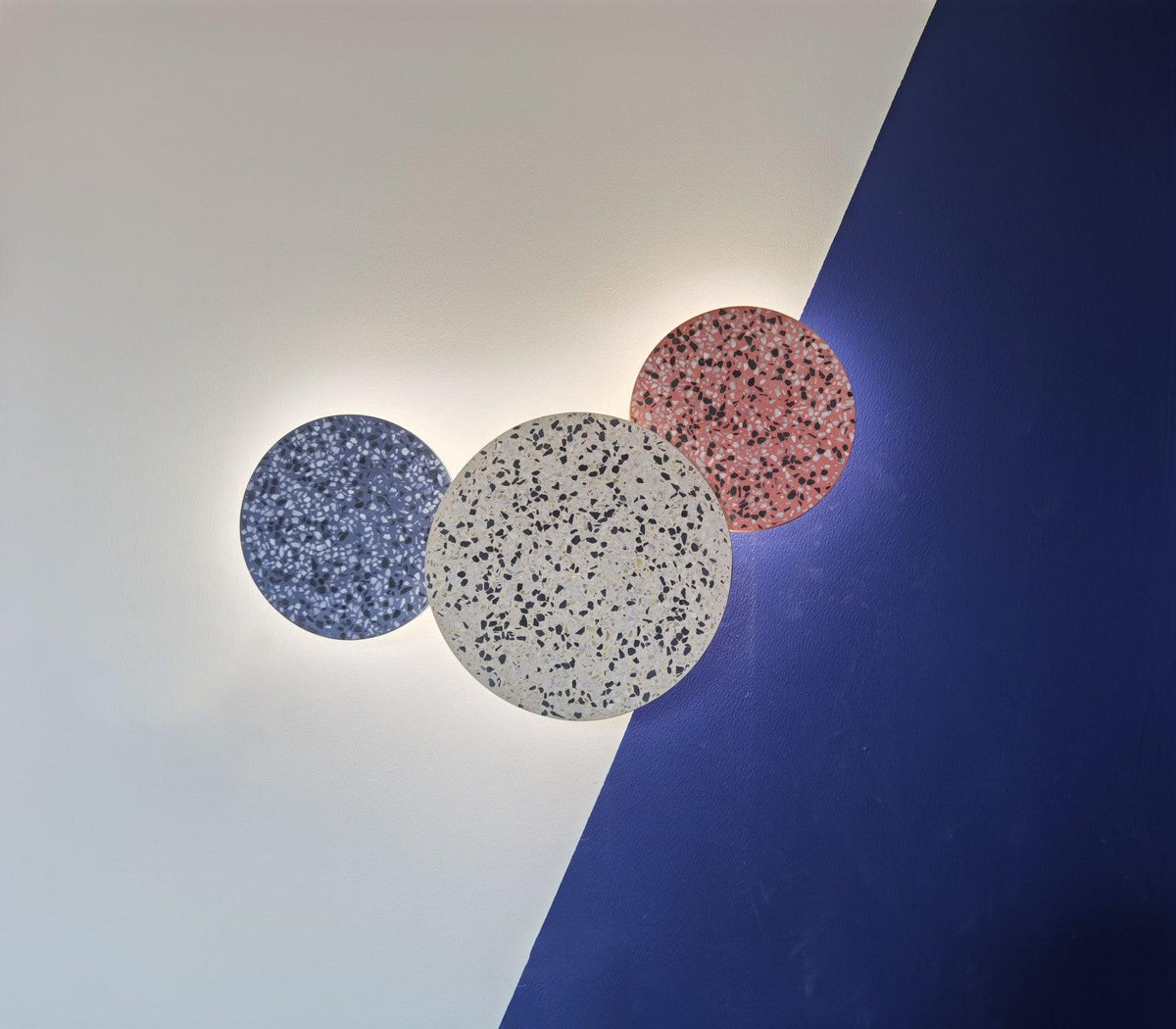 Three Clérachic round wall lights in blue, white and pink terrazzo glow on a wall split into white and deep blue.