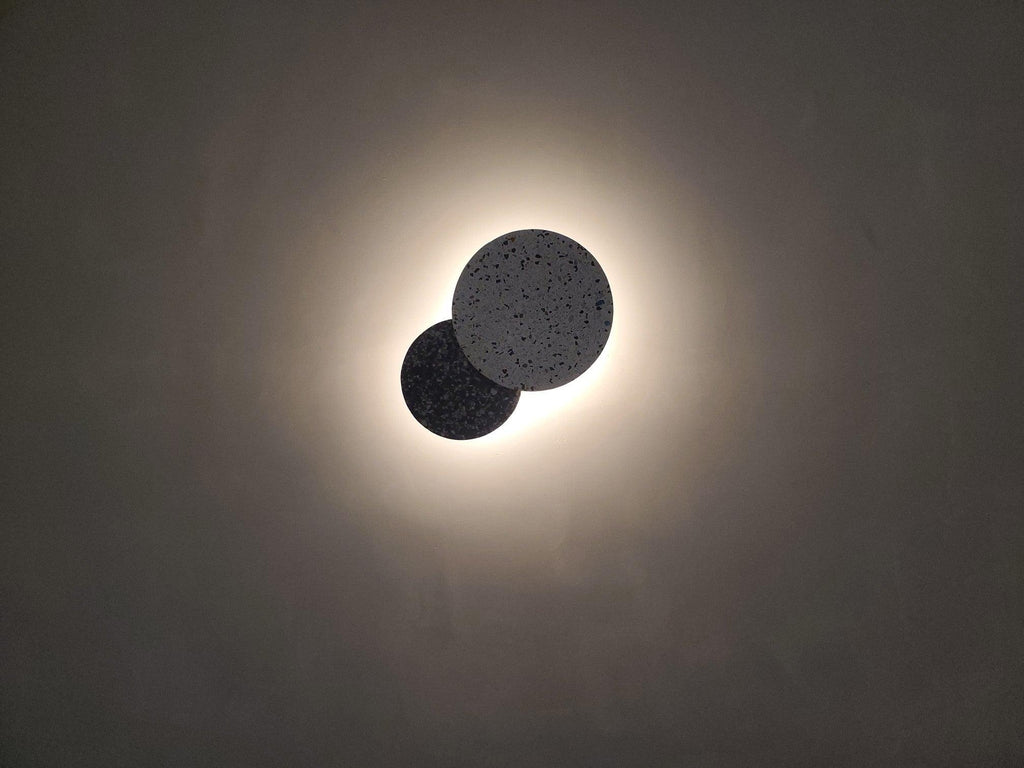 Two overlapping Clérachic round wall lights in black terrazzo give off a soft white glow on a grey wall.