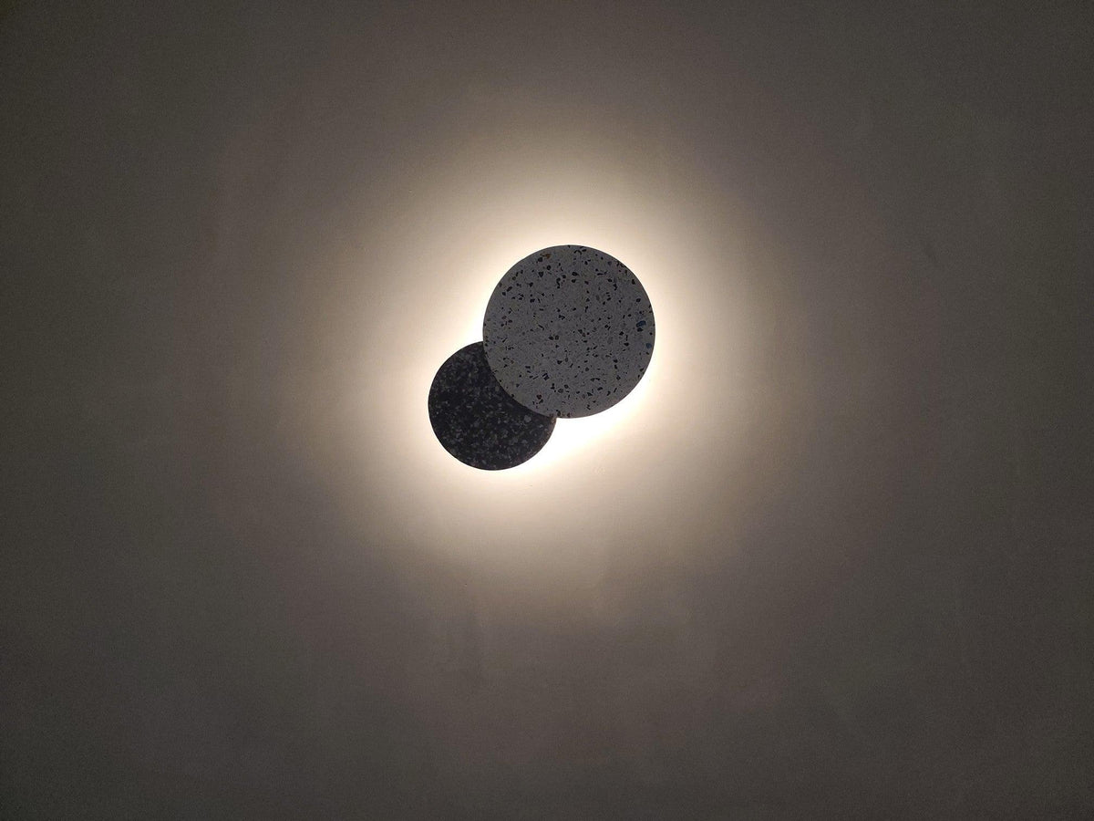 Two overlapping Clérachic round wall lights in black terrazzo give off a soft white glow on a grey wall.