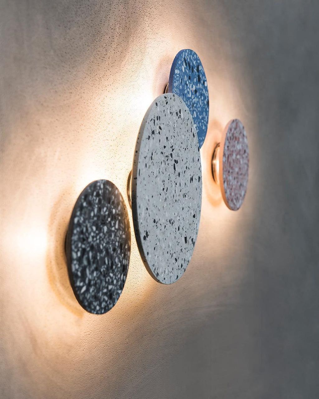 Angled view of three terrazzo Clérachic round wall lights, in black, white and blue, glowing warmly on a grey wall.