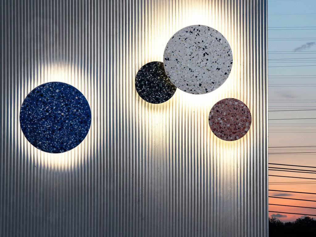 Four Clérachic round wall lights in blue, black, white and pink terrazzo glow on a ribbed metal wall at dusk.
